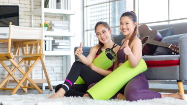İki Milenyum Asyalı genç, sağlıklı, sağlıklı, güçlü, seksi kadın spor modelleri gündelik ürünler, spor kıyafetleri içinde yere oturmuş gülümsüyor yoga minderlerini bir arada tutarak oturma odasında kanepede gitarla birlikte oturuyorlar..