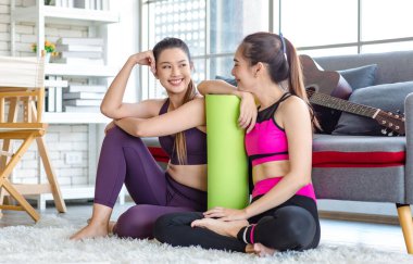 İki Milenyum Asyalı genç, sağlıklı, sağlıklı, güçlü, seksi kadın spor modelleri gündelik ürünler, spor kıyafetleri içinde yere oturmuş gülümsüyor yoga minderlerini bir arada tutarak oturma odasında kanepede gitarla birlikte oturuyorlar..