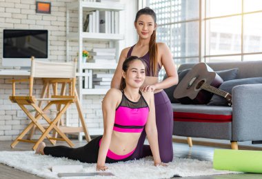 Asyalı, genç, sağlıklı, güçlü, seksi, spor hocası, spor kıyafetleri içinde. Oturma odasındaki özel sınıftaki halı zemininde gerinen öğrencilere yoga öğretmeye yardım ediyor..