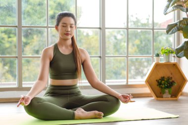 Asyalı, genç, güzel, seksi, zayıf, güçlü spor model. Ekinler içinde, üst üste spor giyimli, bacak bacak üstüne atmış, Lotus pozisyonunda meditasyon yapan, hafif ışık efektiyle sınıfın spor salonundaki yoga minderi..