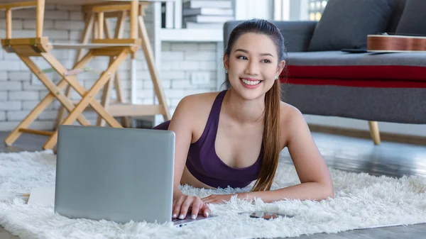 Milenyum Asyalı genç, sağlıklı, sağlıklı, güçlü, seksi kadın spor model. Günlük ürünler, spor kıyafetleri içinde. Oturma odasında halıya uzanmış gülümsüyor ve dizüstü bilgisayardan yoga dersini öğreniyor..