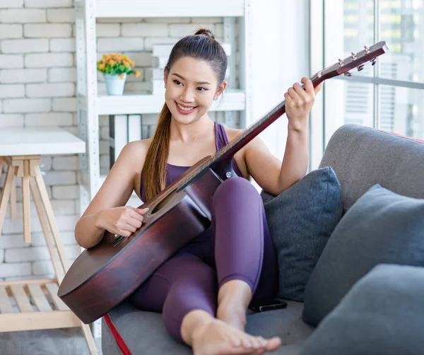 Milenyum Asyalı genç, sağlıklı, güzel, sağlıklı, güçlü, seksi, spor model, rahat giyinmiş, güler yüzlü, evdeki oturma odasındaki rahat koltukta akustik gitar çalan kadın..