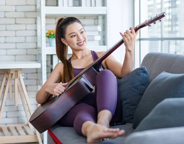 Milenyum Asyalı genç, sağlıklı, güzel, sağlıklı, güçlü, seksi, spor model, rahat giyinmiş, güler yüzlü, evdeki oturma odasındaki rahat koltukta akustik gitar çalan kadın..