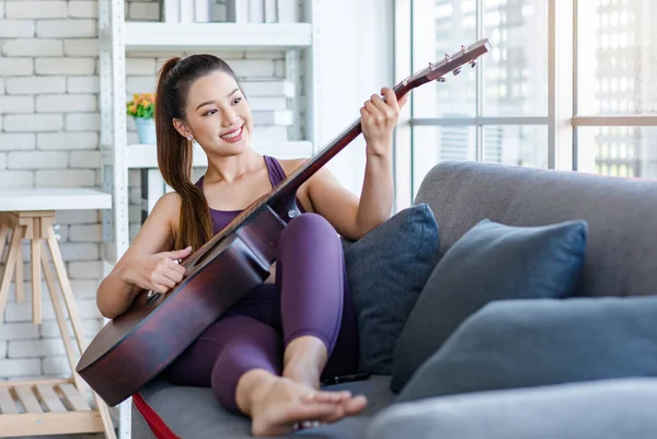 Milenyum Asyalı genç, sağlıklı, güzel, sağlıklı, güçlü, seksi, spor model, rahat giyinmiş, güler yüzlü, evdeki oturma odasındaki rahat koltukta akustik gitar çalan kadın..