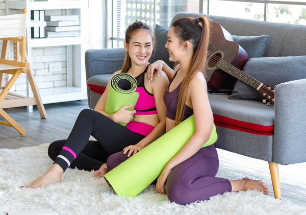 İki Milenyum Asyalı genç, sağlıklı, sağlıklı, güçlü, seksi kadın spor modelleri gündelik ürünler, spor kıyafetleri içinde yere oturmuş gülümsüyor yoga minderlerini bir arada tutarak oturma odasında kanepede gitarla birlikte oturuyorlar..