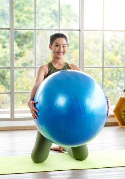 Milenyum Asyalı genç, sağlıklı, sağlıklı, güçlü, seksi kadın spor model, günlük kıyafetleri içinde, güler yüzlü, yoga minderinde egzersiz topuna el ele tutuşmuş, stüdyoda çalışmaya hazır..