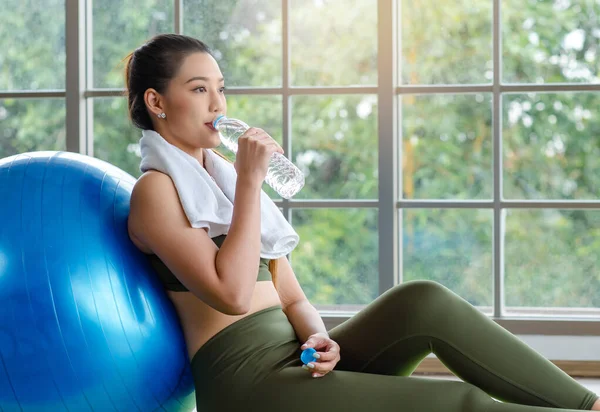 Yakın plan, Millennial Asyalı genç, sağlıklı, güçlü, seksi, spor model, spor kıyafetleri içinde, dinleniyor, egzersiz topuna yaslanıyor, şişede su içiyor..