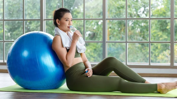 Yakın plan, Millennial Asyalı genç, sağlıklı, güçlü, seksi, spor model, spor kıyafetleri içinde, dinleniyor, egzersiz topuna yaslanıyor, şişede su içiyor..