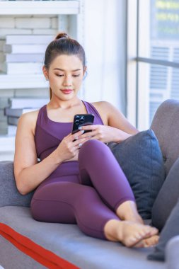 Portre, Millennial Asyalı genç, sağlıklı, sağlıklı, güçlü, seksi kadın spor modelini spor kıyafetleri içinde salonda rahat bir kanepeye uzanmış kameraya gülümseyen bir şekilde, akıllı telefonu çevrimiçi kullanarak çekti..