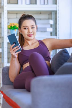 Portre, Millennial Asyalı genç, sağlıklı, sağlıklı, güçlü, seksi kadın spor modelini spor kıyafetleri içinde salonda rahat bir kanepeye uzanmış kameraya gülümseyen bir şekilde, akıllı telefonu çevrimiçi kullanarak çekti..