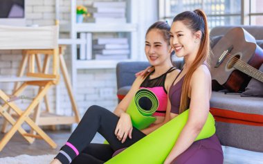 İki Milenyum Asyalı genç, sağlıklı, sağlıklı, güçlü, seksi kadın spor modelleri gündelik ürünler, spor kıyafetleri içinde yere oturmuş gülümsüyor yoga minderlerini bir arada tutarak oturma odasında kanepede gitarla birlikte oturuyorlar..