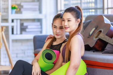 İki Milenyum Asyalı genç, sağlıklı, sağlıklı, güçlü, seksi kadın spor modelleri gündelik ürünler, spor kıyafetleri içinde yere oturmuş gülümsüyor yoga minderlerini bir arada tutarak oturma odasında kanepede gitarla birlikte oturuyorlar..