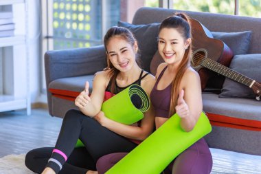 İki Milenyum Asyalı genç, sağlıklı, sağlıklı, güçlü, seksi kadın spor modelleri gündelik ürünler, spor kıyafetleri içinde yere oturmuş gülümsüyor yoga minderlerini bir arada tutarak oturma odasında kanepede gitarla birlikte oturuyorlar..