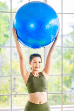 Milenyum Asyalı genç, sağlıklı, sağlıklı, güçlü, seksi kadın spor model, günlük kıyafetleri içinde, güler yüzlü, yoga minderinde egzersiz topuna el ele tutuşmuş, stüdyoda çalışmaya hazır..