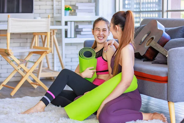 İki Milenyum Asyalı genç, sağlıklı, sağlıklı, güçlü, seksi kadın spor modelleri gündelik ürünler, spor kıyafetleri içinde yere oturmuş gülümsüyor yoga minderlerini bir arada tutarak oturma odasında kanepede gitarla birlikte oturuyorlar..