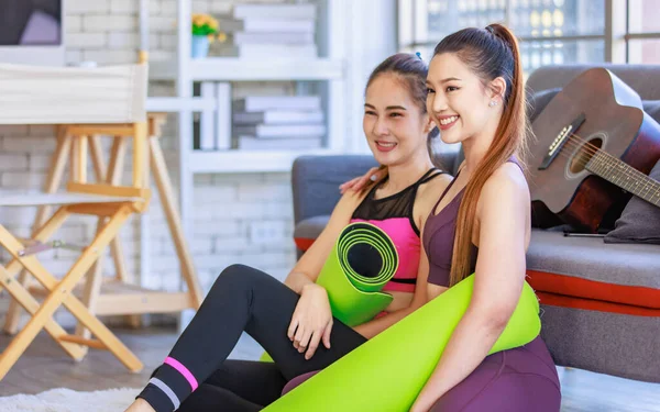 İki Milenyum Asyalı genç, sağlıklı, sağlıklı, güçlü, seksi kadın spor modelleri gündelik ürünler, spor kıyafetleri içinde yere oturmuş gülümsüyor yoga minderlerini bir arada tutarak oturma odasında kanepede gitarla birlikte oturuyorlar..