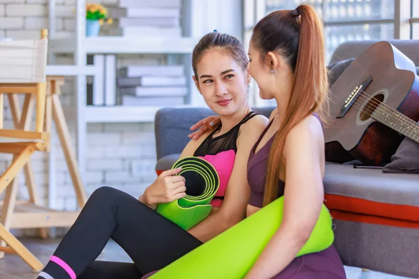 İki Milenyum Asyalı genç, sağlıklı, sağlıklı, güçlü, seksi kadın spor modelleri gündelik ürünler, spor kıyafetleri içinde yere oturmuş gülümsüyor yoga minderlerini bir arada tutarak oturma odasında kanepede gitarla birlikte oturuyorlar..
