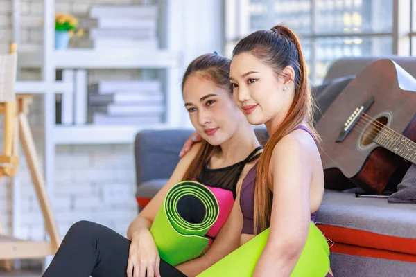 İki Milenyum Asyalı genç, sağlıklı, sağlıklı, güçlü, seksi kadın spor modelleri gündelik ürünler, spor kıyafetleri içinde yere oturmuş gülümsüyor yoga minderlerini bir arada tutarak oturma odasında kanepede gitarla birlikte oturuyorlar..