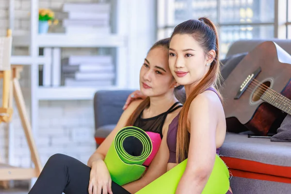 İki Milenyum Asyalı genç, sağlıklı, sağlıklı, güçlü, seksi kadın spor modelleri gündelik ürünler, spor kıyafetleri içinde yere oturmuş gülümsüyor yoga minderlerini bir arada tutarak oturma odasında kanepede gitarla birlikte oturuyorlar..