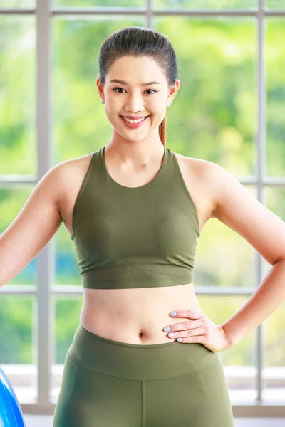 Millennial Asian young healthy slim strong sexy female sporty model in casual crop top sportswear sitting smiling posing holding hand on exercise ball on yoga mat ready to workout in classroom studio.