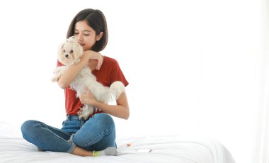 Asyalı, genç, mutlu, kısa saçlı kız modelinin stüdyo fotoğrafı. Günlük kıyafet giymiş, bacak bacak üstüne atmış, gülümseyen, kucaklaşan, beyaz arka planda uzun tüylü, yavru köpek yavrusu Malta köpeği taşıyan..