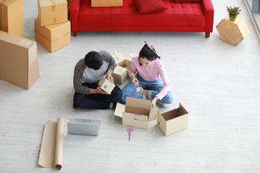 Asyalı genç iş adamı dizüstü bilgisayarda oturup müşteriyle sohbet ediyor. Serbest çalışan kadın işkadını, çevrimiçi mağazada satılan karton kutularda ürün paketlemeye yardım ediyor..