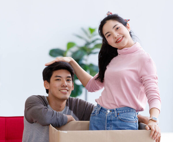 Asian young lovely lover couple female girlfriend surprise male boyfriend sitting on red sofa in living room at home by hiding inside big cardboard box as present gift for his Birthday celebration.