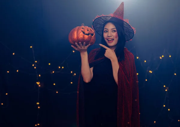 Studio shot of Asian young beautiful teenager girl wearing Halloween costume with tall witch hat black rope and orange dress standing smiling holding blue cocktail glass drink in trick & treat party.