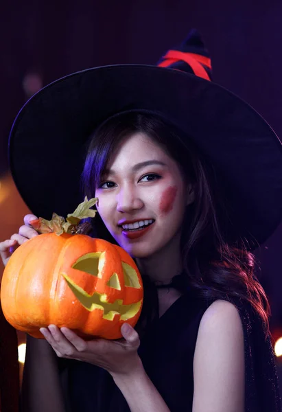 Studio shot of Asian young beautiful teenager girl wearing Halloween costume with tall witch hat black rope and orange dress standing smiling holding blue cocktail glass drink in trick & treat party.