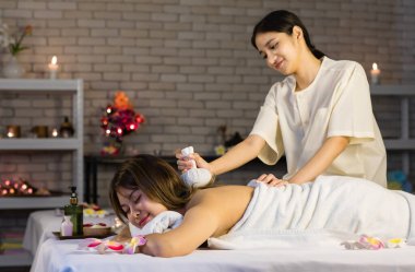 Asyalı, genç, seksi, neşeli kadın spa müşterisi beyaz temiz banyo havlusuyla kaplanmış yatarken tanınmayan masöz sıcak bitkisel masaj ve sırt masajı kullanarak gülümsüyor..