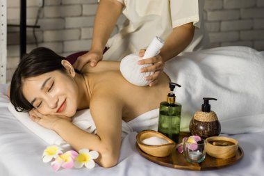 Asyalı, genç, seksi, neşeli kadın spa müşterisi beyaz temiz banyo havlusuyla kaplanmış yatarken tanınmayan masöz sıcak bitkisel masaj ve sırt masajı kullanarak gülümsüyor..