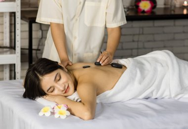 Asyalı genç ve güzel bir spa müşterisi masaj yatağının üzerine uzanmış gülümserken tanınmayan profesyonel kadın masajcı sırt omurgasına masaj yaparak masaj yapıyor..