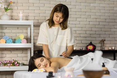 Asyalı genç ve güzel bir spa müşterisi masaj yatağının üzerine uzanmış gülümserken tanınmayan profesyonel kadın masajcı sırt omurgasına masaj yaparak masaj yapıyor..