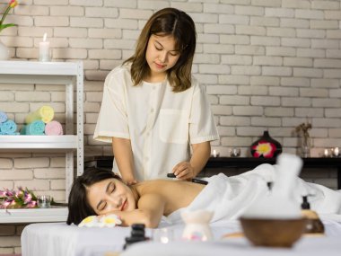 Asyalı genç ve güzel bir spa müşterisi masaj yatağının üzerine uzanmış gülümserken tanınmayan profesyonel kadın masajcı sırt omurgasına masaj yaparak masaj yapıyor..