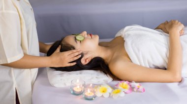 Asyalı, genç, güzel bir spa müşterisi masaj yatağında organik salatalıkla kaplı gözlerini kapatırken tanınmayan profesyonel kadın masaj terapisti kafa ve yüz masajı yapıyor..