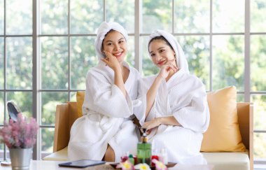 İki Asyalı genç ve güzel spa müşterisi, beyaz bornozlu, kafaları havluyla kaplı, gülümseyen, aromaterapi masaj seansından sonra sağlıklı yanaklara ve yüzlere dokunan arkadaşlar..