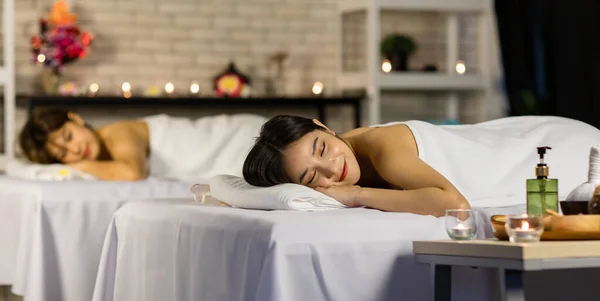 Closeup shot Asian young sexy female spa customer covered by white clean bath towel lay down on massaging bed with flowers smiling look at camera while girlfriend lying beside in blurred background.