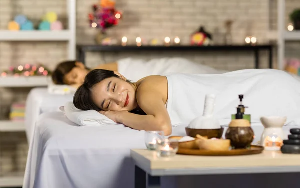 Closeup shot Asian young sexy female spa customer covered by white clean bath towel lay down on massaging bed with flowers smiling look at camera while girlfriend lying beside in blurred background.