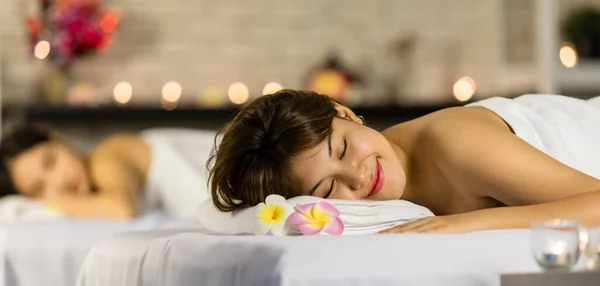 Closeup shot Asian young sexy female spa customer covered by white clean bath towel lay down on massaging bed with flowers smiling look at camera while girlfriend lying beside in blurred background.