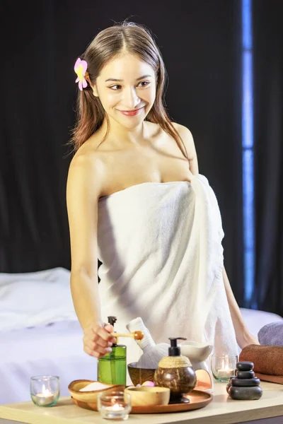 Asian young beautiful sexy female spa customer in white clean bath towel standing behind massage cosmetic table select aromatherapy perfume oil bottle smiling look at camera.