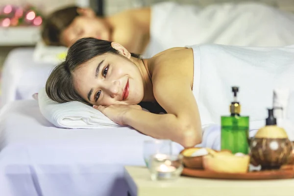 Closeup shot Asian young sexy female spa customer covered by white clean bath towel lay down on massaging bed with flowers smiling look at camera while girlfriend lying beside in blurred background.