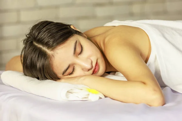 Closeup Asian young beautiful sexy happy female spa customer covered by white clean bath towel laying down resting relaxing closed eyes smiling on massaging bed putting bloom orchid flower on ear.