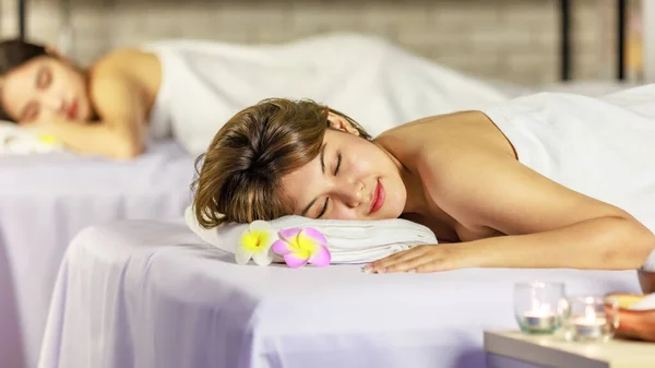 Closeup shot Asian young sexy female spa customer covered by white clean bath towel lay down on massaging bed with flowers smiling look at camera while girlfriend lying beside in blurred background.