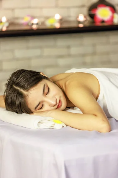 Closeup Asian young beautiful sexy happy female spa customer covered by white clean bath towel laying down resting relaxing closed eyes smiling on massaging bed putting bloom orchid flower on ear.