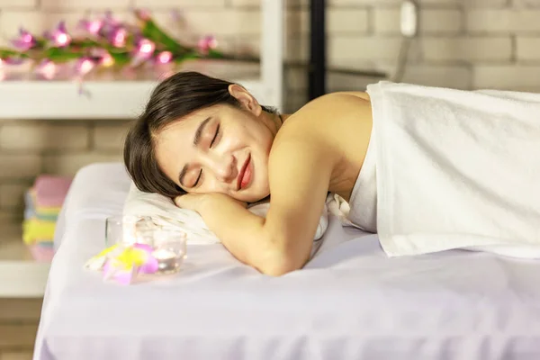 Closeup Asian young beautiful sexy happy female spa customer covered by white clean bath towel laying down resting relaxing closed eyes smiling on massaging bed putting bloom orchid flower on ear.