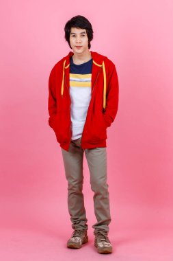 Asyalı genç şehirli erkek modelin, sokak tarzı giyinmiş kırmızı ceketli, pantolonun cebinde el ele poz veren fotoğrafının tüm vücut resmi. Pembe arka plandaki kameraya bakın..