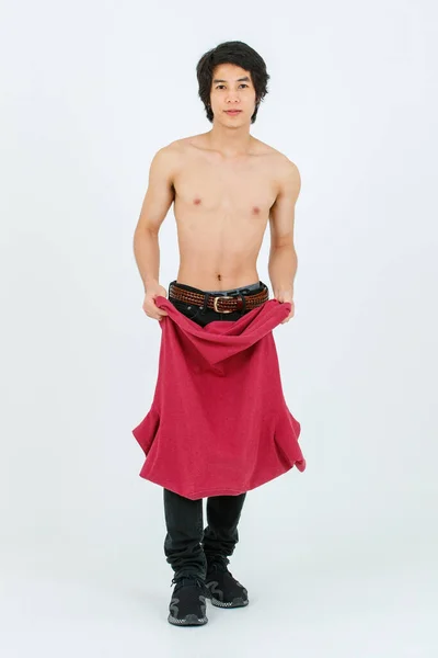 ortrait isolated cutout full body studio shot of Asian young handsome confident slim shirtless teenager male model standing holding red t shirt in hands showing muscular torso on white background.