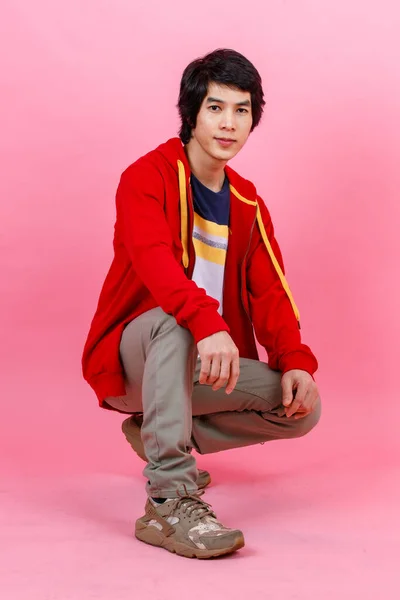 Portrait full body studio shot of Asian young urban teenager fashion male model in street style outfit red jacket sitting posing look at camera on pink background.