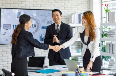Two happy millennial professional successful businesswomen in formal suit standing shaking hands greeting together when business deal agreement done while businessman entrepreneur smiling clapping.