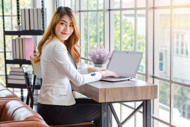 Asyalı genç, düşünceli, profesyonel, başarılı, beyaz takım elbiseli kadın iş kadını tezgahta oturup ofis binasının dışına bakıp dizüstü bilgisayar kullanarak daktilo yazmayı düşünüyor..